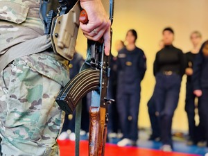 zdjęcie przedstawia uczniów klasy mundurowej i policyjnego instruktora trzymającego karabinek AKMS w ręku