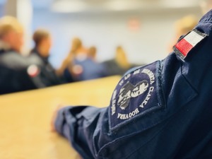 zdjęcie przedstawia uczniów klasy mundurowej siedzących przy stolikach, na pierwszym planie rękaw munduru z naszywką szkoły