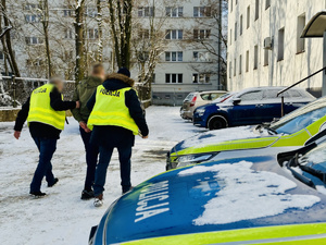 Nieumundurowani policjanci w kamizelkach z napisem Policja prowadzą osobę zatrzymaną