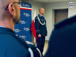 zdjęcie z sali odpraw pokazujące komendanta stojącego na środku sali, w pierwszym planie ramiona policjantów skierowanych twarzą w stronę przemawiającego