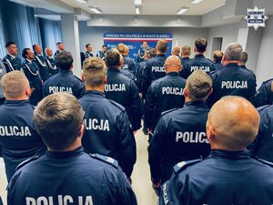 zdjęcie z sali odpraw pokazujące grupę policjantów stojących w rzędach, w tle widoczny przemawiający komendant