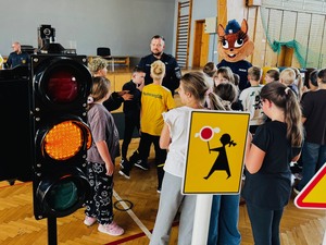 sygnalizator świetlny, obok znaki drogowe i stojący przy policjancie uczniowie